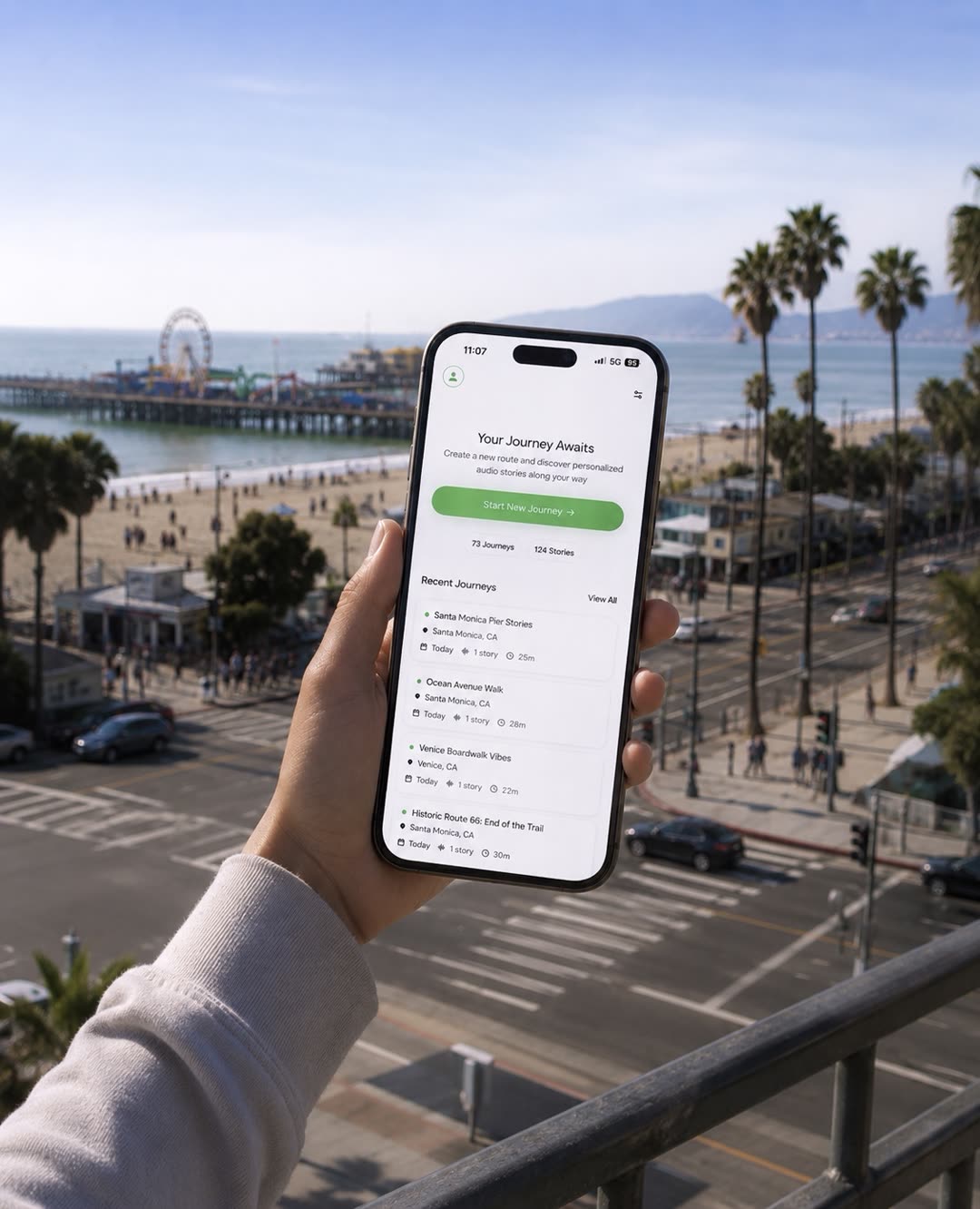 A hand holding a phone above the Santa Monica Pier, showing the Guroute app's journey list including Santa Monica Pier Stories, Ocean Avenue Walk, Venice Boardwalk Vibes, and Historic Route 66.